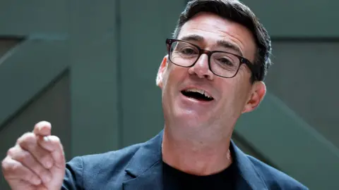 Andy Burnham speaking and smiling in front of a green door wearing a dark suit and glasses
