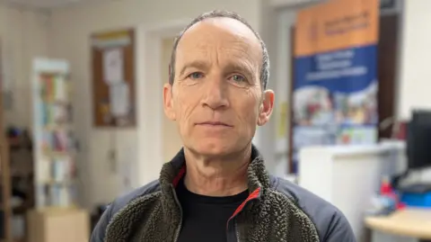 A man in a black T-shirt and dark fleece is looking into the camera. He is sat inside the South West MS Therapy Centre. 