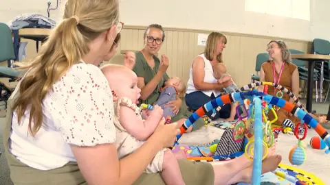 A group of women are sitting on the floor with their babies at a MotherBorn session. Three mothers are talking to each other and another mother is talking to co-founder, Steph Pope. There are toys scattered across the floor for the children to use.