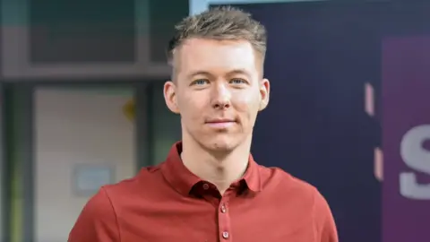 Nick Sheridan has brown hair and wears a red shirt and smiles at the camera. He is presenting a BBC TV show.