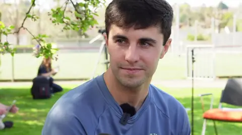 Liam Walsh has dark brown short hair and is wearing a blue T-shirt with a fluffy black mic attached. He is looking off to the side of the frame and is sitting in front of a sports field.