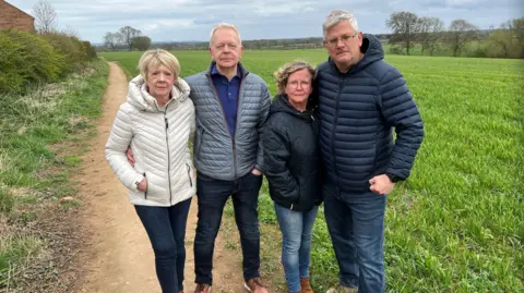This picture shows four members of the Kirby Hill - Protect Our Village campaign group, who claim plans for dozens of new homes will overload the local sewage and drainage system. The two men and two women are standing in the "greenfield" site developers want to build on. None of them are smiling. There is a long footpath worn into the ground to their left and some trees, still without their leaves, in the middle-distance. 