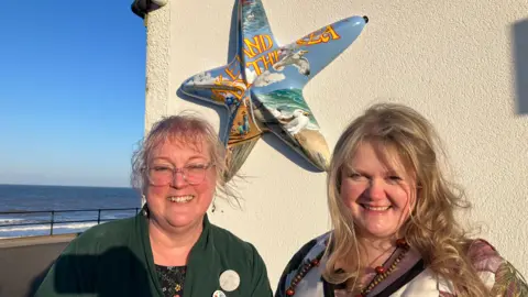 A woman with a blonde fringe and hair tied back is wearing large clear-rimmed glasses and a green cardigan and is smiling into the camera. Next to her is a woman with long blonde hair also smiling into the camera. Behind them is a wall with a starfish sculpture which says 'Lake-Land By The Sea' in yellow lettering with a background of the seaside with seagulls, sky, sand and the sea. The actual sea can be seen behind them.
