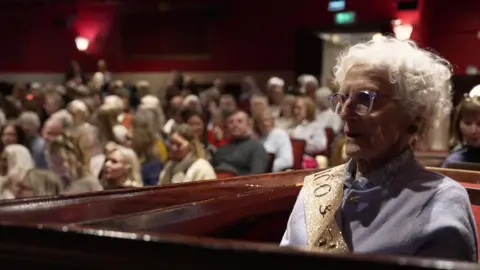 Cilla sits in a box in the theatre audience watching the performance intently.