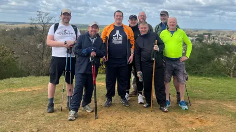 For Men To Talk A group of men, out for a walk, holding walking sticks, with houses, trees and bushes behind them.