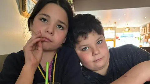 One young boy and one young girl with black hair, grey t-shirts and dark-coloured eyes looking into the camera, with a green straw in front of the girl.