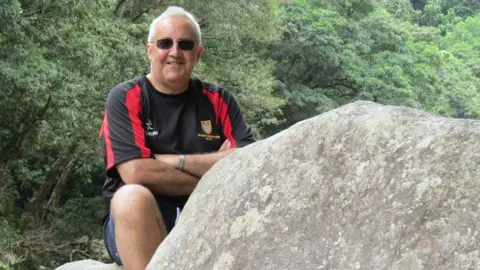 A man sitting outdoors on large rocks, smiling with arms folded, wearing sunglasses and a black and red sports shirt, surrounded by green trees.