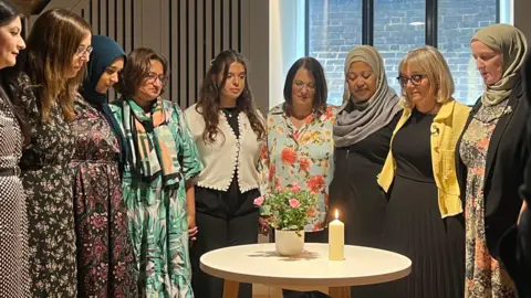 BBC Muslim and six Jewish women standing around a table, looking at a lit candle and a potted miniature rose