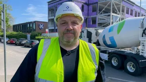 Bob Hockenhull/BBC A man wearing a black T-shirt with a yellow hi-vis jacket over the top. He is standing in front of a building under construction, with a cement mixer parked next to it. He is also wearing a white hard hat, and he has a grey stubbly beard.