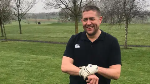 A man wearing a black polo-shirt while smiling at the camera. He is wearing white gloves while holding onto a golf club. He is standing in a field 