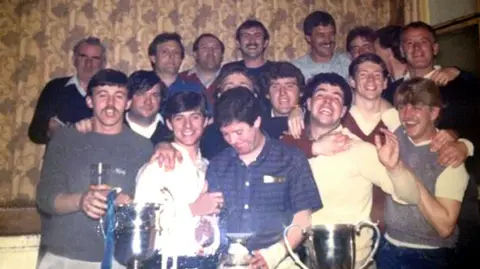 John Burns A group of men are smiling and laughing as they stand in front of three trophies 