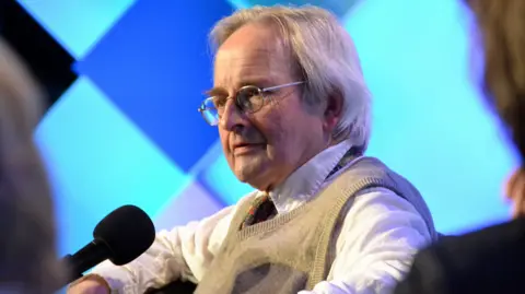 Allan Massie, a man in his 80s with white hair, silver-rimmed spectacles and wearing a white shirt, paisley pattern red tie and beige slip-over, sits in front of a microphone at an event. There is a blue and torquoise patterned background.