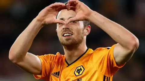 Diogo Jota with short brown hair in a yellow and black Wolves football kit making a heart symbol with his hands and smiling.