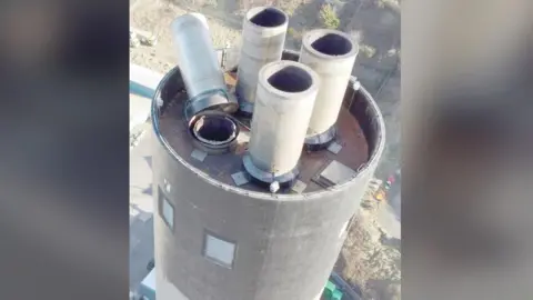 An ariel view of the Peel power station stack, which has four white chimney caps, one of which has fallen over, leaving the inside of the stack exposed.