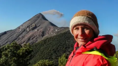 Supplied A photograph of a woman smiling wearing a red jacket and a hat. There is a volcano mountain behind her and blue skies. 