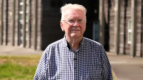 A man with white hair and glasses standing on grass in front of a wooden school building