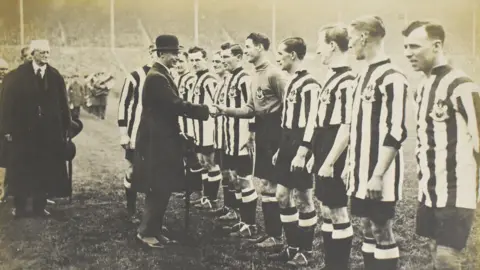 Anderson & Garland Auctioneers Players stand in a line in Newcastle black and white kit. The king is in a bowler hat and shakes one of the player's hand.