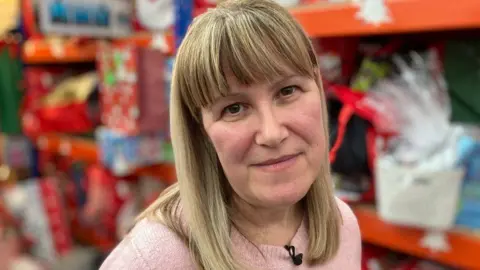 A head shot of Jo Andrew - she has blonde shoulder-length hair and is wearing a pink jumper. Behind her are shelves of Christmas gifts.  