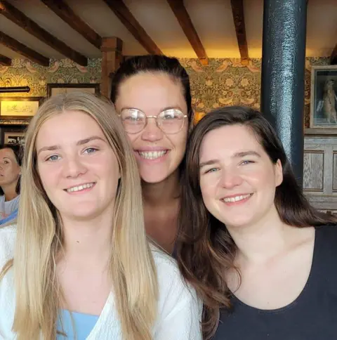 Hunt Family Three young women one with long blonde hair, another with glasses and dark hair pulled back and the third with long brown hair. They are all looking straight into the camera and smiling.