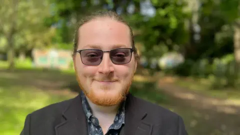 BBC Steven Lewkowicz, 25, wearing tinted glasses, a beard, and hair in a pony tail, smiles at the camera in a park