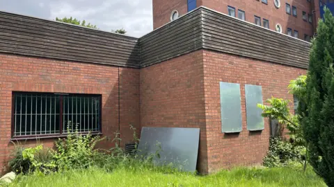 Overgrown grass and shrubs surround a red brick building. A window is visible and it has white bars behind it. A large metal sheet has been propped against a wall in the centre of the image and two other windows on the right have been boarded over.