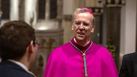 Bishop Nicholas Hudson wearing a pink cassock and hat. He is smiling and looking at a man to the left.