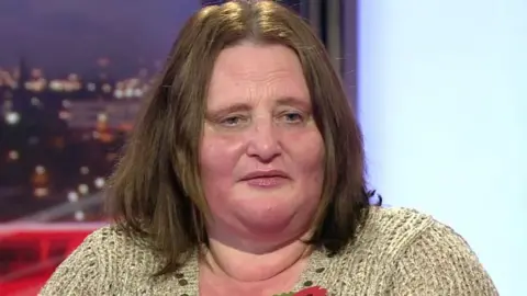 Karen Downes, wearing a poppy and beige cardigan, she speaks in a BBC TV studio.