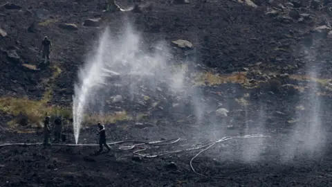 Reuters Image shows firefighters walking on the scorched moorland. Punctured hoses are being used to create sprinkler systems.
