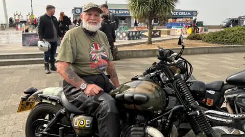 Richard Daniel/BBC Paul Vincent sits on his motorbike. He wears a green cap with a green T-shirt and leather trousers. He has a long grey beard.