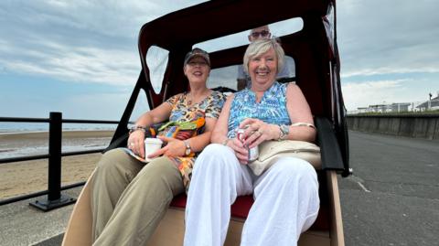 Two women, one wearing a cap, leopard print top and khaki linen trousers and the other with blonde hair wearing a blue top and white trousers, sat in a trishaw buggy on a seafront.