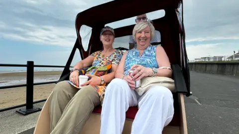 BBC Two women, one wearing a cap, leopard print top and khaki linen trousers and the other with blonde hair wearing a blue top and white trousers, sat in a trishaw buggy on a seafront.