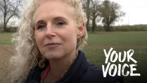 A woman with long, blonde, curly hair smiles at the camera. She is wearing a dark top with a pink collar. Behind her is a green field. 