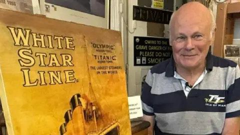 Peter Boyd-Smith stood in his shop wearing a black and grey polo shirt with white hair and is smiling. 