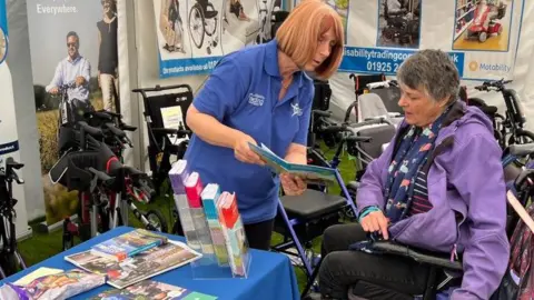 DAD A female volunteer shows a leaflet to a female wheelchair user in a marquee containing a number of mobility walkers and scooters