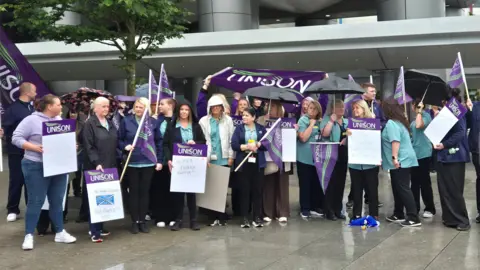 A group of hospital workers, holding signs and banners, outside a large hospital building 