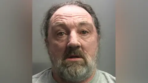 Cumbria Police A mugshot of James McGlinchey. He is wearing a light grey sweater and he has a grey beard with wiry grey hair.