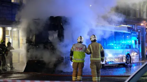 Zara Lazarov A smoking bus sits at the side of a road. The back end is burnt out and blackened, but the fire is mostly out. Firefighters standing on the pavement on the left direct a jet of water towards the bus and two firefightersin the foreground look on at the wreck.
