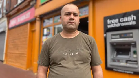 A man in front of an orange shop wearing a light brown T-shirt