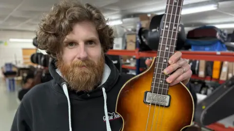 A picture of Luke Hobbs, a man in his 30s with curly, light brown hair and reddish beard, holding up a Hofner bass and looking into camera.