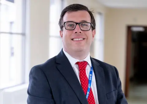 Queen Elizabeth Hospital, King's Lynn Chief operating officer, Simon Illingworth looking at the camera. The picture shows his head and shoulders and he is wearing black glasses. He has a red tie and a black hacket on. He is smiling and standing in a hospital corridor