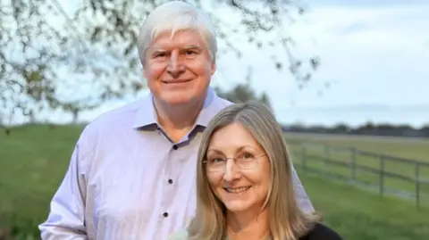 University of Cambridge A man with white/grey hair and wearing a pale blue shirt stands next to a shorter woman with blonde hair and glasses