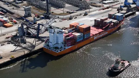 PA Media A orange container ship is berthed alongside a long concrete quay at a port. A crane which appears bent and broken is lying on top of some of the containers. Some of the containers also appear damaged. The river is a dark brown with glints of sunlight reflecting off ripples of water.
