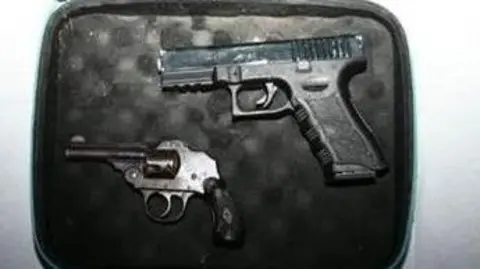 Two guns - a Glock-style pistol and a small vintage-looking enclosed-hammer revolver - lie in a small case which has black padding.