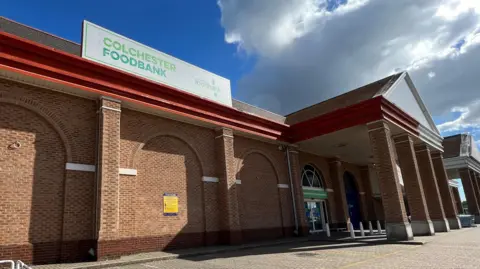 Richard Smith/BBC Colchester Foodbank is a large brick warehouse building at a retail park. It has tall columns out the front and a white sign advertising its name on top.