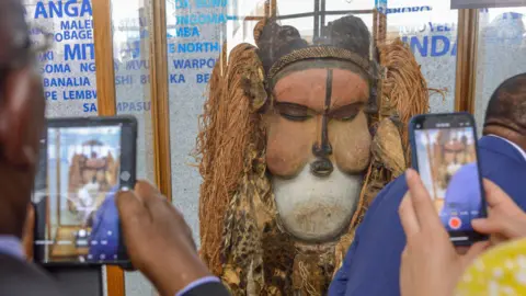 AFP People take photos of the Kakuungu mask after its unveiling in Kinshasa, DR Congo - Wednesday 8 June 2022