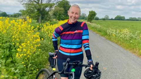 Julie smiling to the camera on a country lane with green fields, trees and cow parsley in the background. She is wearing a striped blue and mauve cycling jacket and is leaning against her black bike with a helmet in her hand.