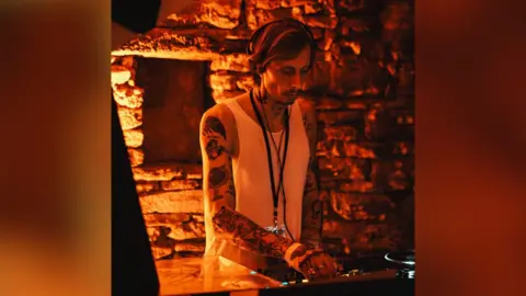 Steve Stone Steve is djing at a club. The venue has orange lighting. He is wearing a vest and has multiple tattoos on both his arms.