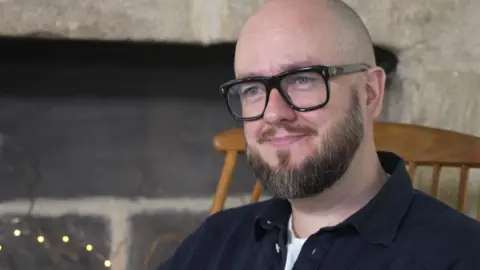 A man with a bald head and a brown beard and glasses, wearing a navy shirt over a white t shirt. He is sitting in a wooden chair and smiling. 