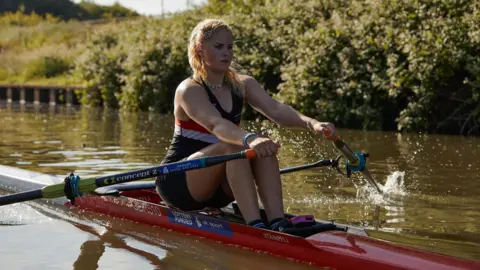 Hartpury College A female single skull Hartpury College rower on a red skull. SHe is wearing a rowing vest and shorts and has an oar in each hand, while on the river with trees behind her.