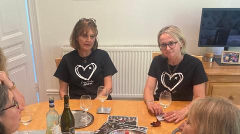 A darker haired woman and a fair haired women wearing black t-shirts saying Cambridge Widows sitting at a table